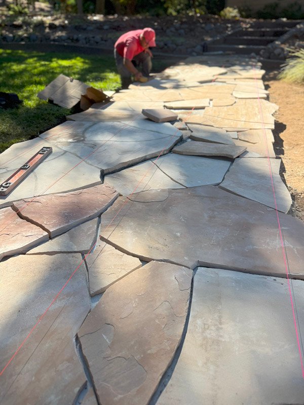 Workers from South Canyon Construction carefully place large stones to create a stunning path in a bright garden. The sunny day helps them see each piece clearly as they work.